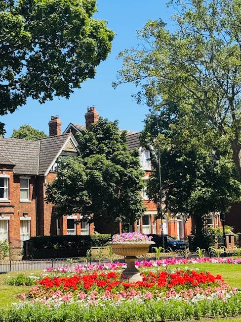 A well-maintained urban garden situated in front of a row of historic brick residential buildings under a clear blue sky. The garden features a central stone planter filled with vibrant pink and purpl
