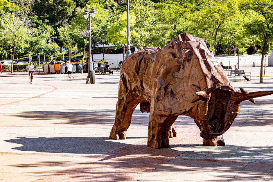 A large wooden sculpture of a rhinoceros is displayed in an open urban plaza surrounded by mature trees with lush green leaves. The sculpture, made from roughly hewn wooden panels, showcases detailed